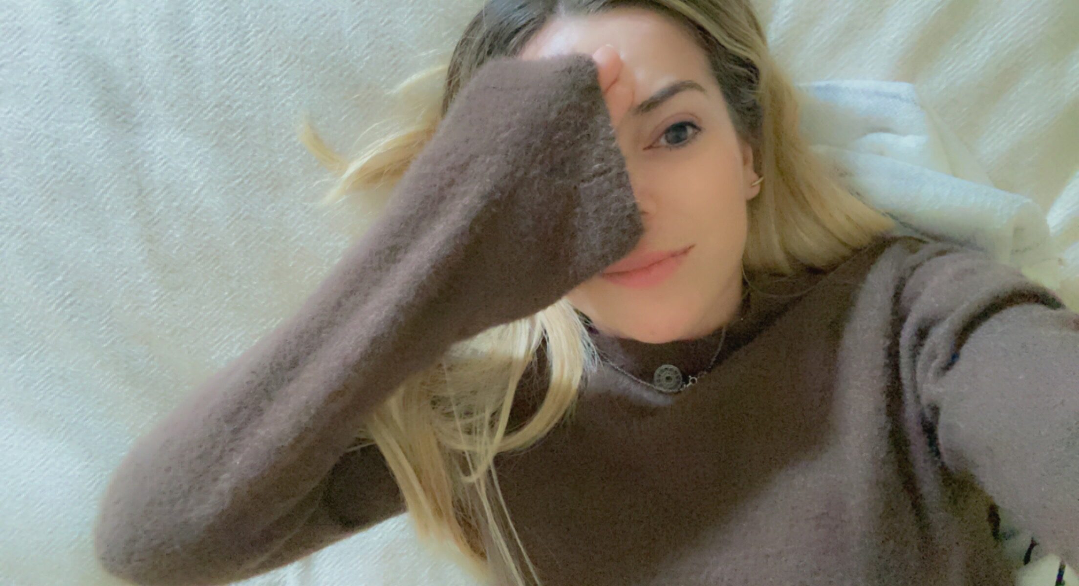 Imagem de mimi do MimiBlog deitada sobre a cama com uma camisola castanha a esboçar um sorriso e com uma mão virada do avesso a tapar metade da cara